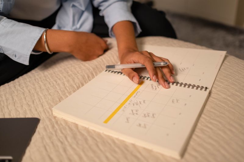 Two hands over an open spiral-bound planner, a pen in one of them, one lane of its grid marked out in yellow highlighter