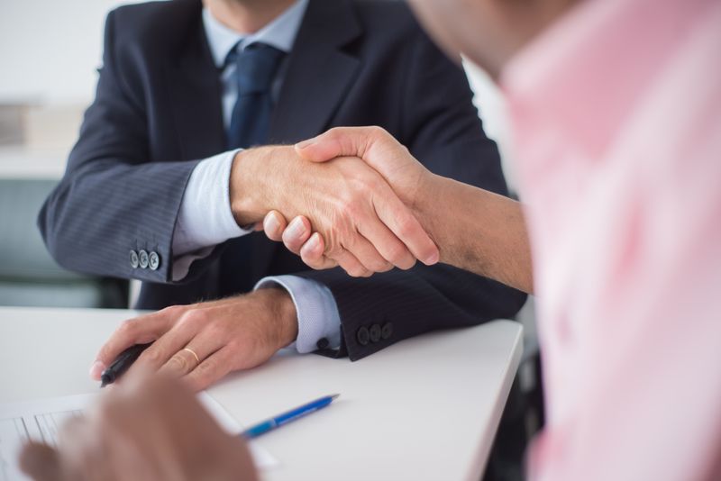 Two people shaking hands across a table beside a document and pen
