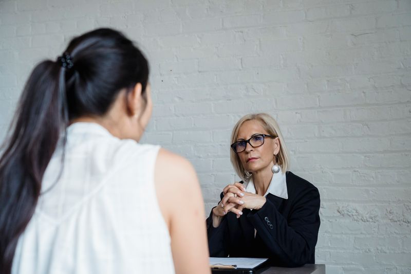 Woman in a black blazer and glasses listening with her hands clasped, facing a colleague seen from behind, against a white painted brick wall
